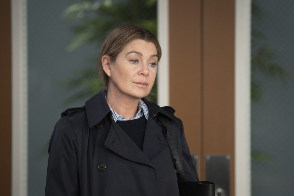 Ellen Pompeo standing in a hospital office in a black coat as Dr Meredith Grey in Grey's Anatomy.