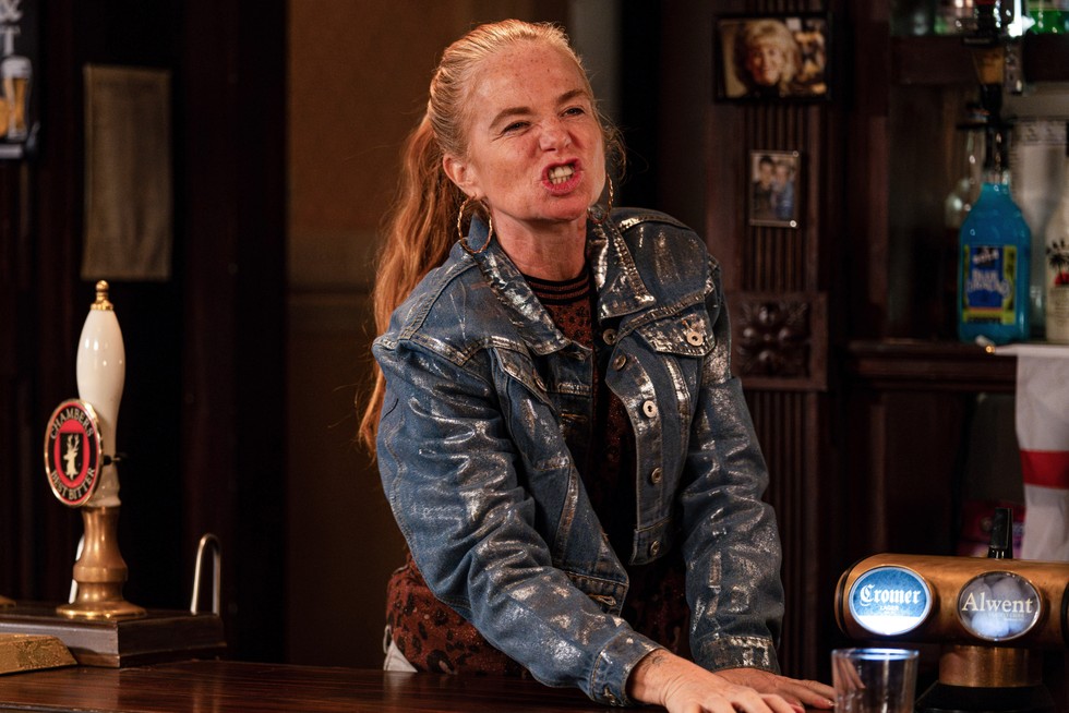 Patsy Palmer as Bianca Jackson standing behind the bar of the Queen Vic with an angry face mid-speech westing a shiny jacket and her hair tied back in a long pony tail in EastEnders.