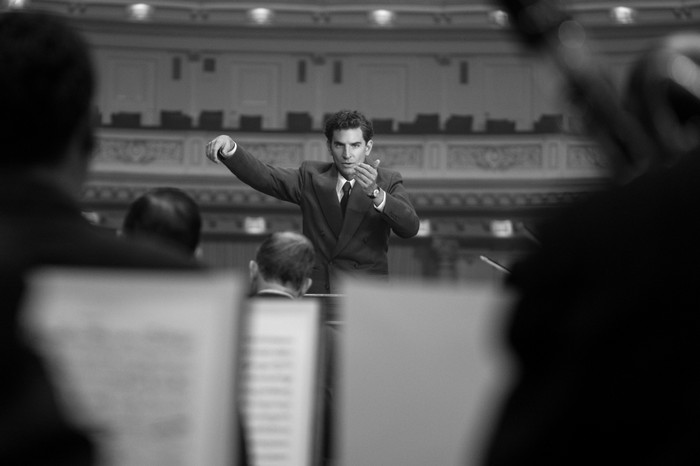 Bradley Cooper as Leonard Bernstein in Maestro wearing a suit, conducting an orchestra