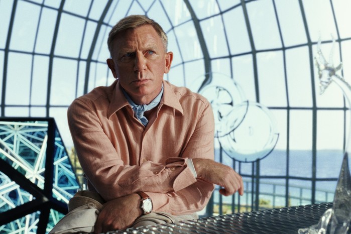 Daniel Craig as Benoit Blanc in Glass Onion in an orange shirt sat in a glass building