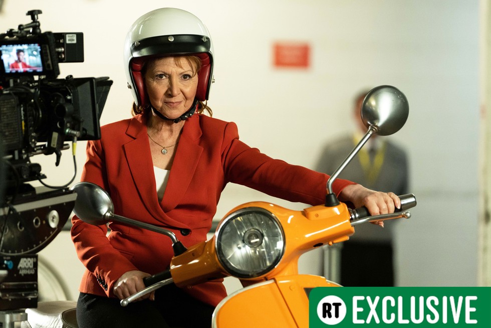 Bonnie Langford sits atop a yellow Vespa with a white helmet on and red blazer in a shot from filming Doctor Who in Doctor Who Unleashed. The RT Exclusive sticker sits in the bottom right corner of the image.