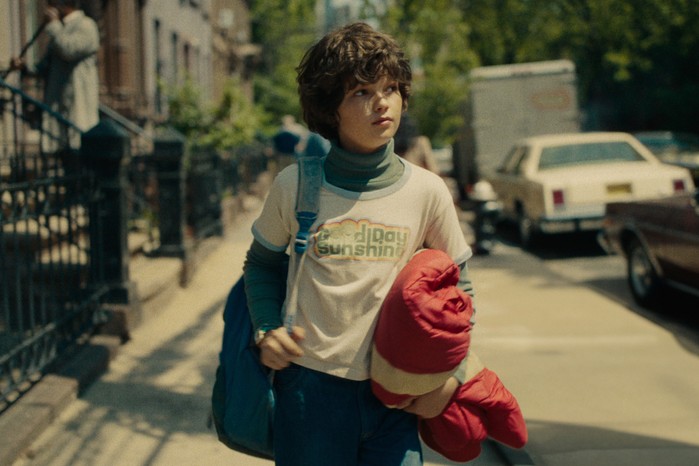 Ivan Howe as Edgar in Eric wearing a Good Day Sunshine t-shirt and walking down a street holding a red jacket and blue bag.