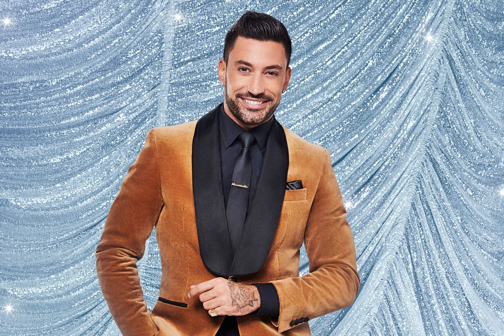 Giovanni Pernice smiling in front of a blue curtain background. He is wearing a brown velvet suit with one hand in front of him and the other in his pocket.