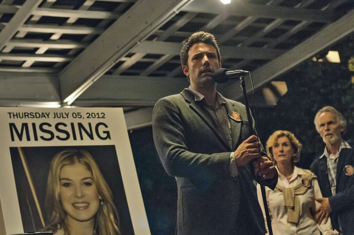 Ben Affleck as Nick Dunne in Gone Girl, stood by a missing poster featuring Amy Elliot Dunne, as played by Rosamund Pike