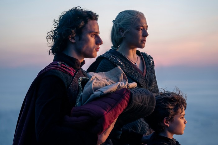 Harry Collett as Prince Jacaerys Velaryon holding a baby, Emma D'Arcy as Queen Rhaenyra Targaryen and Oscar Eskinkanzi as Prince Joffrey Velaryon stare out to sea in House of the Dragon season 2.