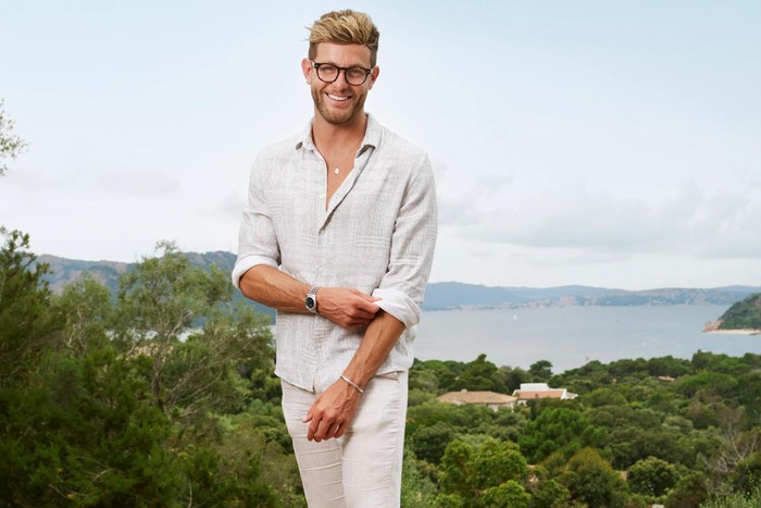 Harvey Armstrong wearing a cream outfit, with trees and water in the background