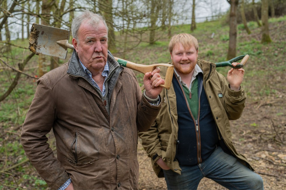 Jeremy Clarkson and Kaleb Cooper stood in the middle of a woodland looking ahead. They are both wearing farming clothing and have shovels resting on their shoulders.