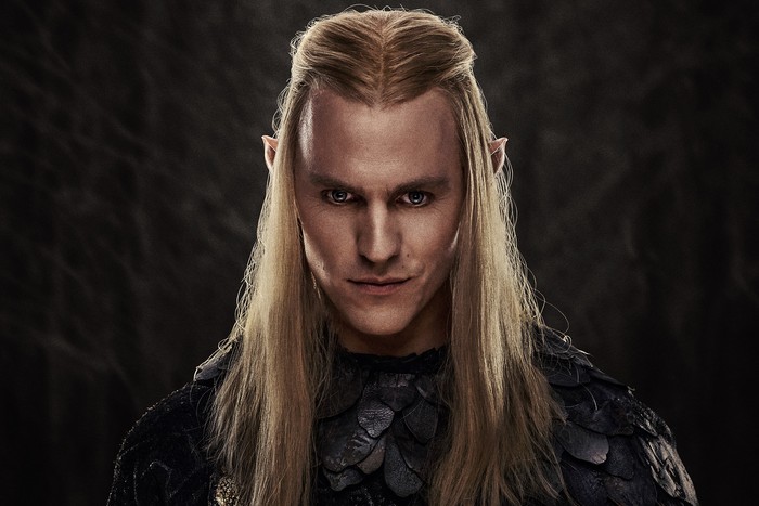 Charlie Vickers as Sauron standing and looking into the camera wearing a black suit with long blonde hair.