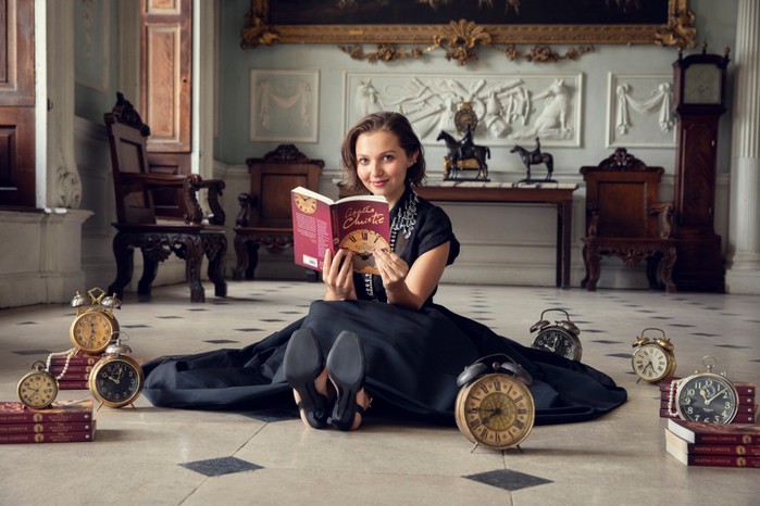 Mia McKenna-Bruce reading The Seven Dials Mystery and sat on the floor with clocks all around her