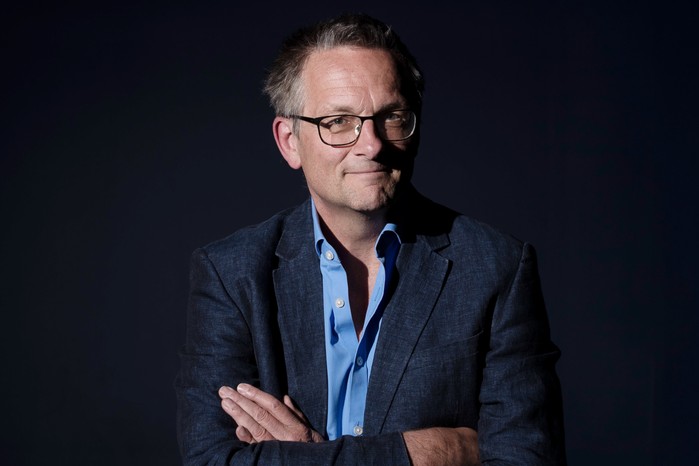 Dr Michael Mosley with his arms crossed, looking at the camera in front of a dark black background.