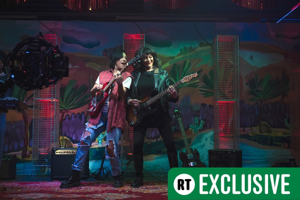 Sarah Kameela Impey as Saira and Meera Syal as Sister Squire in We Are Lady Parts season 2. They are on stage playing guitar and there is a green banner in the bottom right corner reading RT Exclusive