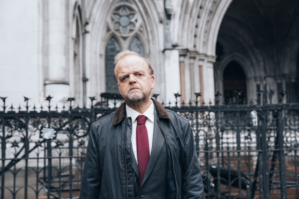 Alan Bates (played by Toby Jones) stands outside a courthouse with a serious facial expression