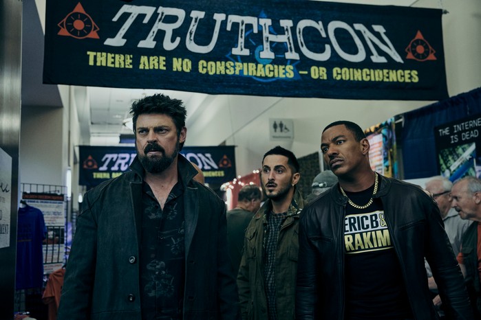 Karl Urban as Billy Butcher, Laz Alonso as Mother's Milk and Tomer Capone as Frenchie in The Boys, stood under a banner which says Truthcon