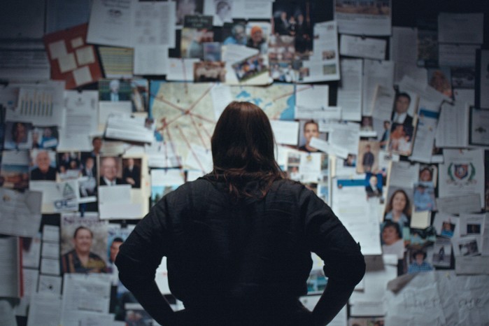 Katherine Kubler standing with her back to the camera and hands on her hips standing in front of a board filled with evidence and photos in The Program: Cons, Cults and Kidnapping.