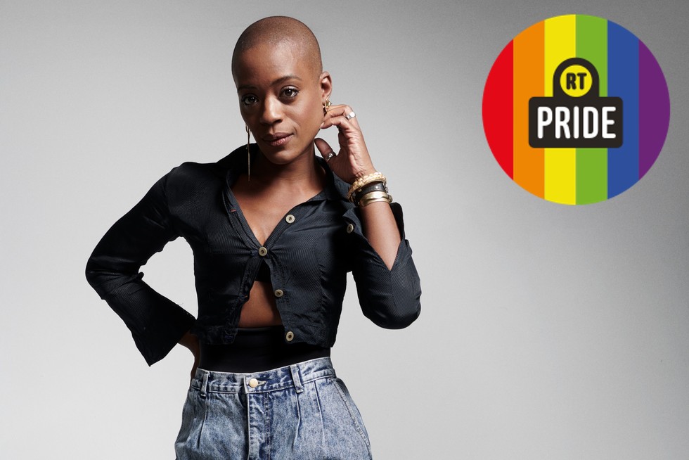 T'Nia Miller looking at the camera standing against a white background. She's wearing jeans and a black top. In the upper right corner, a rainbow badge reads: RT Pride.